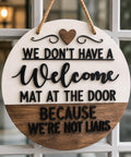 Round white-and-wood door hanger with raised black lettering reading “We don’t have a welcome mat at the door because we’re not liars,” a wooden heart, and a rope hanger.