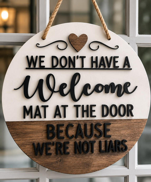 Round white-and-wood door hanger with raised black lettering reading “We don’t have a welcome mat at the door because we’re not liars,” a wooden heart, and a rope hanger.
